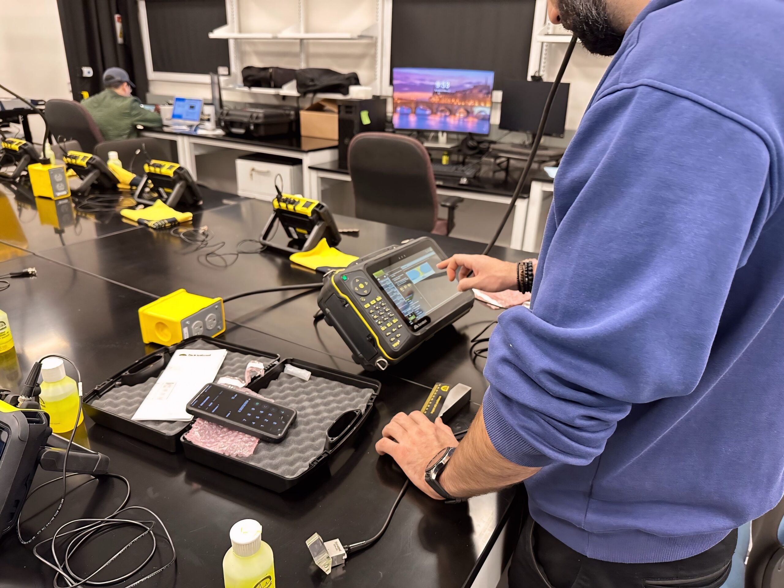 A man in a blue sweatshirt stands at a lab table using a handheld electronic testing device with a touchscreen display. The device is connected by a cable to a small sensor placed on the table. Nearby are a smartphone, a foam-lined equipment case, and a small bottle of yellow liquid. Additional yellow testing units sit on the table, and computer workstations and shelves are visible in the background.