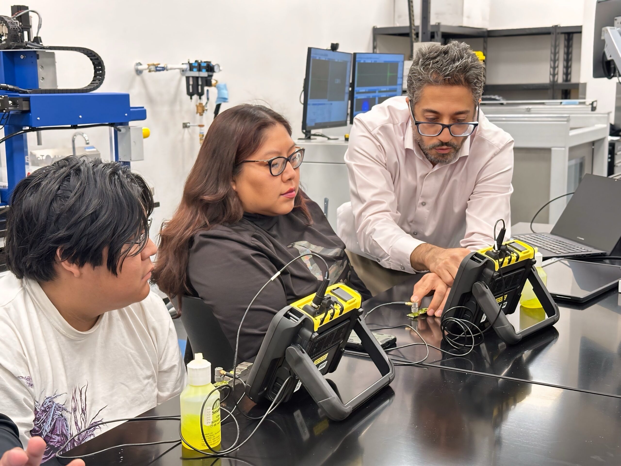 Three people sit and stand around a lab table in a technical workspace. An instructor in a light-colored shirt points to a small component connected to a yellow handheld testing device, while two students watch closely. The devices are propped up on stands and connected by cables. A small bottle of yellow liquid and a laptop sit on the table, with industrial equipment and computer monitors visible in the background.