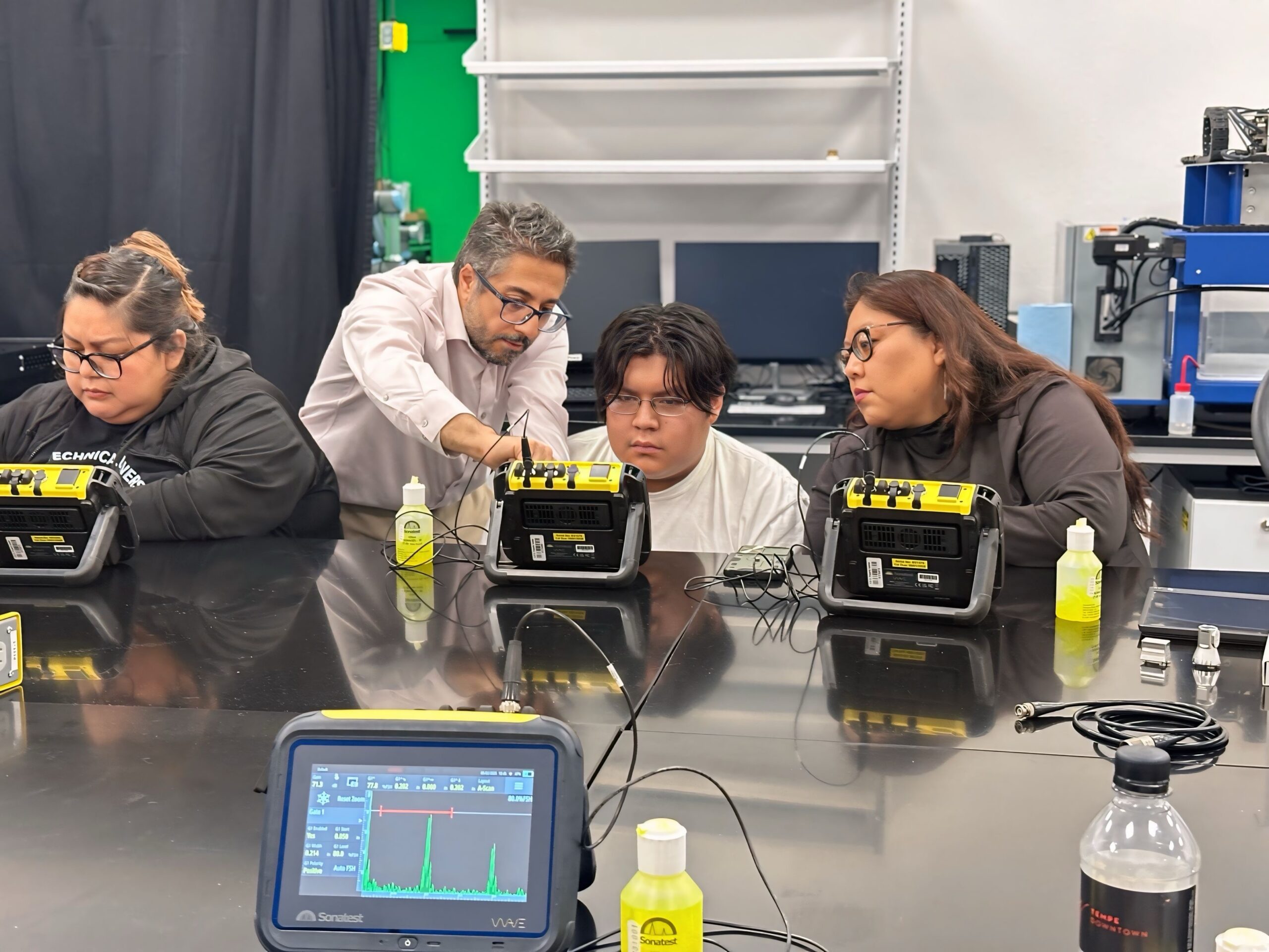 Four people sit and stand around a reflective lab table, each with a yellow handheld testing device propped on a stand. An instructor leans in to adjust a probe connected to one device while a student watches closely. Two other students focus on their own instruments. A monitor in the foreground displays a graph with peaks, and small bottles of yellow liquid, cables, and lab equipment are arranged across the table.